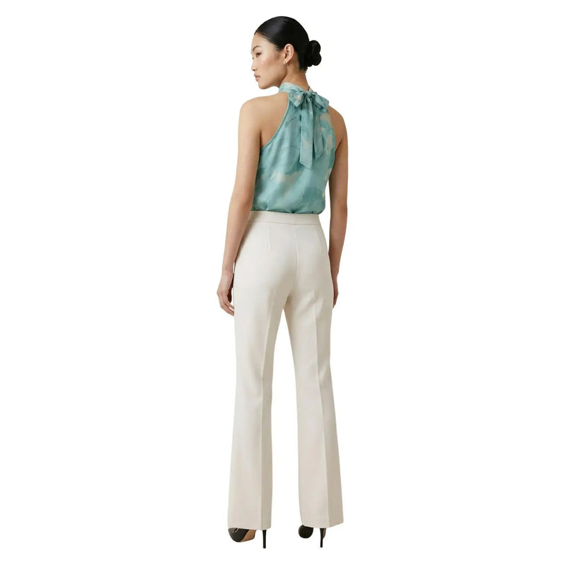 A woman wearing Artigli women trousers white zip front pockets in cream with a teal silk halter top with a bow at the back
