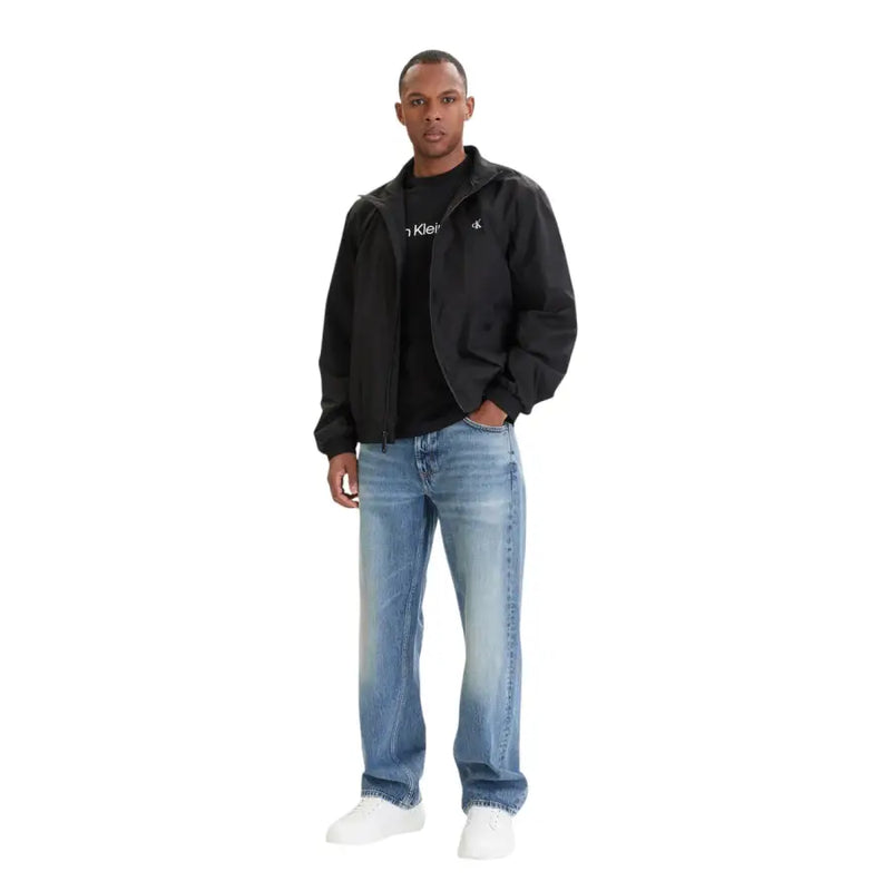Schwarz Calvin Klein Jacke mit dezenten Logos auf Brust und Ärmel, getragen über passendem schwarzem t-shirt mit blauen Jeans aus 100 % Baumwolle