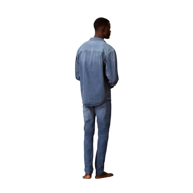 Un homme portant un jean Calvin Klein bleu en coton, coupe slim, et une chemise en jean délavée assortie.