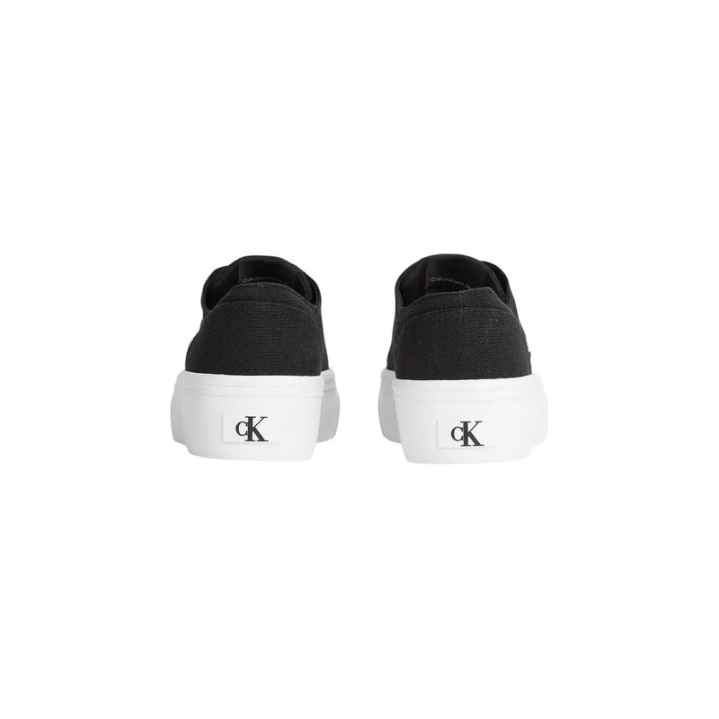 Black canvas slip-on sneakers with thick white rubber sole and ck logo on heel shown from the back side