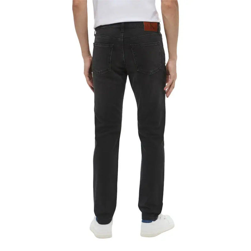 Back view of dark wash black denim jeans by calvin klein featuring a brown leather brand patch on the waistband