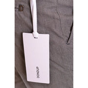 White rectangular brand tag with dondup printed in black attached by white ribbon to grey houndstooth fabric of men’s cotton trousers