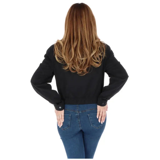 Woman with long wavy brown hair wearing a black blazer and blue jeans from behind