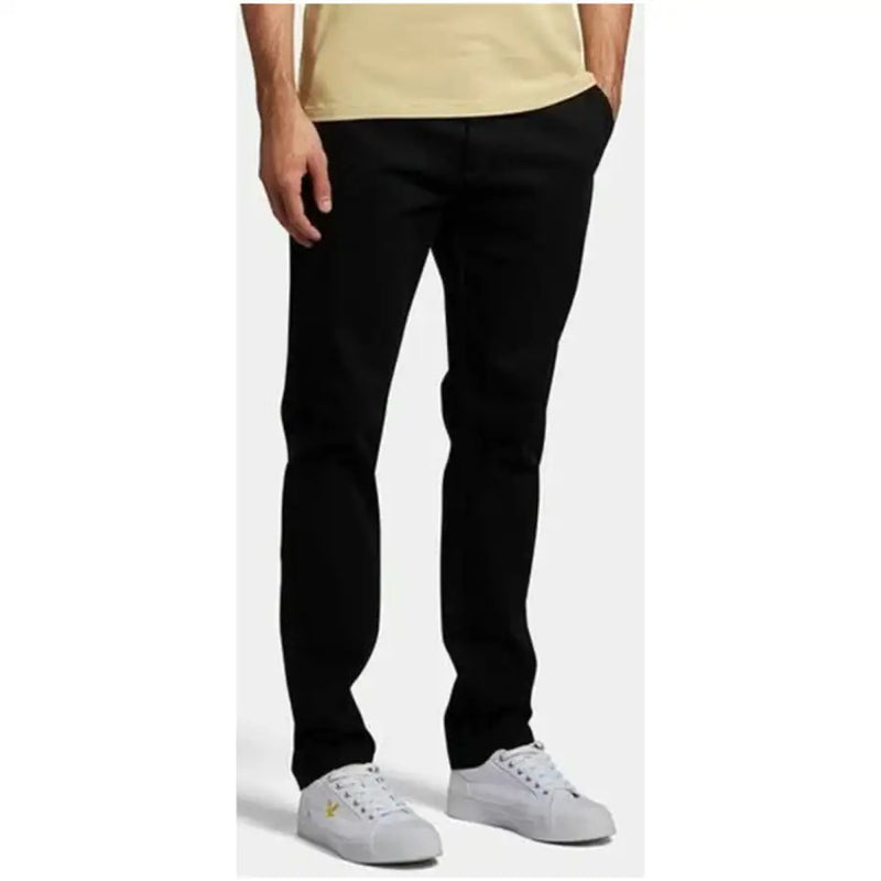 Lyle & Scott Schwarze, schmal geschnittene Herrenhose aus Baumwollmischgewebe mit dezentem goldenem Logo, kombiniert mit weißen Sneakers.
