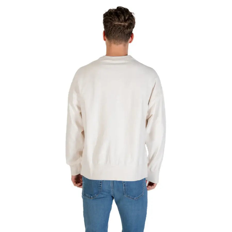 Man wearing a beige crewneck sweatshirt and blue jeans facing sideways