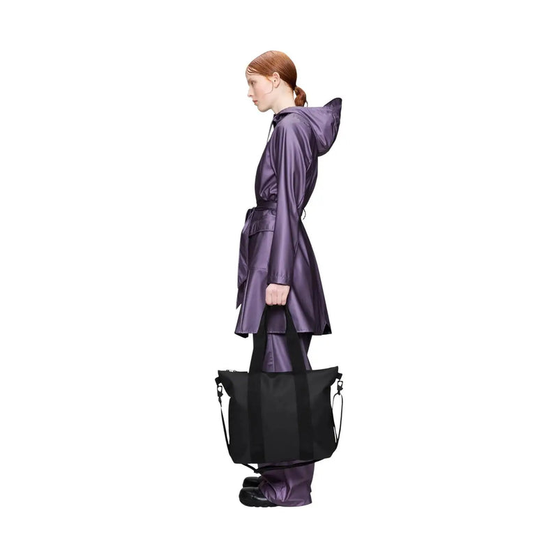 A sleek black Rains women tote bag mini in glossy polyester with sturdy handles held by a model in a purple raincoat