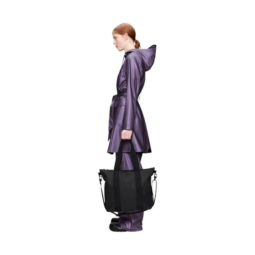 A sleek black Rains women tote bag mini in glossy polyester with sturdy handles held by a model in a purple raincoat