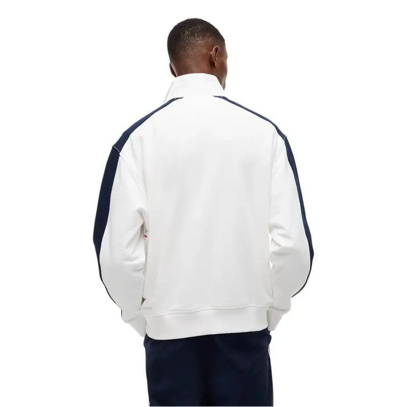 Veste de survêtement Lacoste blanche avec empiècements contrastants bleu marine aux manches et aux épaules, portée avec un jean Tommy Hilfiger et un sweat-shirt en coton bio à rayures blanches.